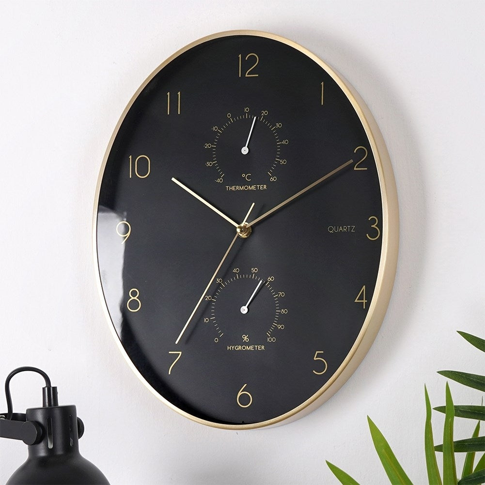 Un reloj de pared negro con marco dorado, con termómetro e higrómetro.