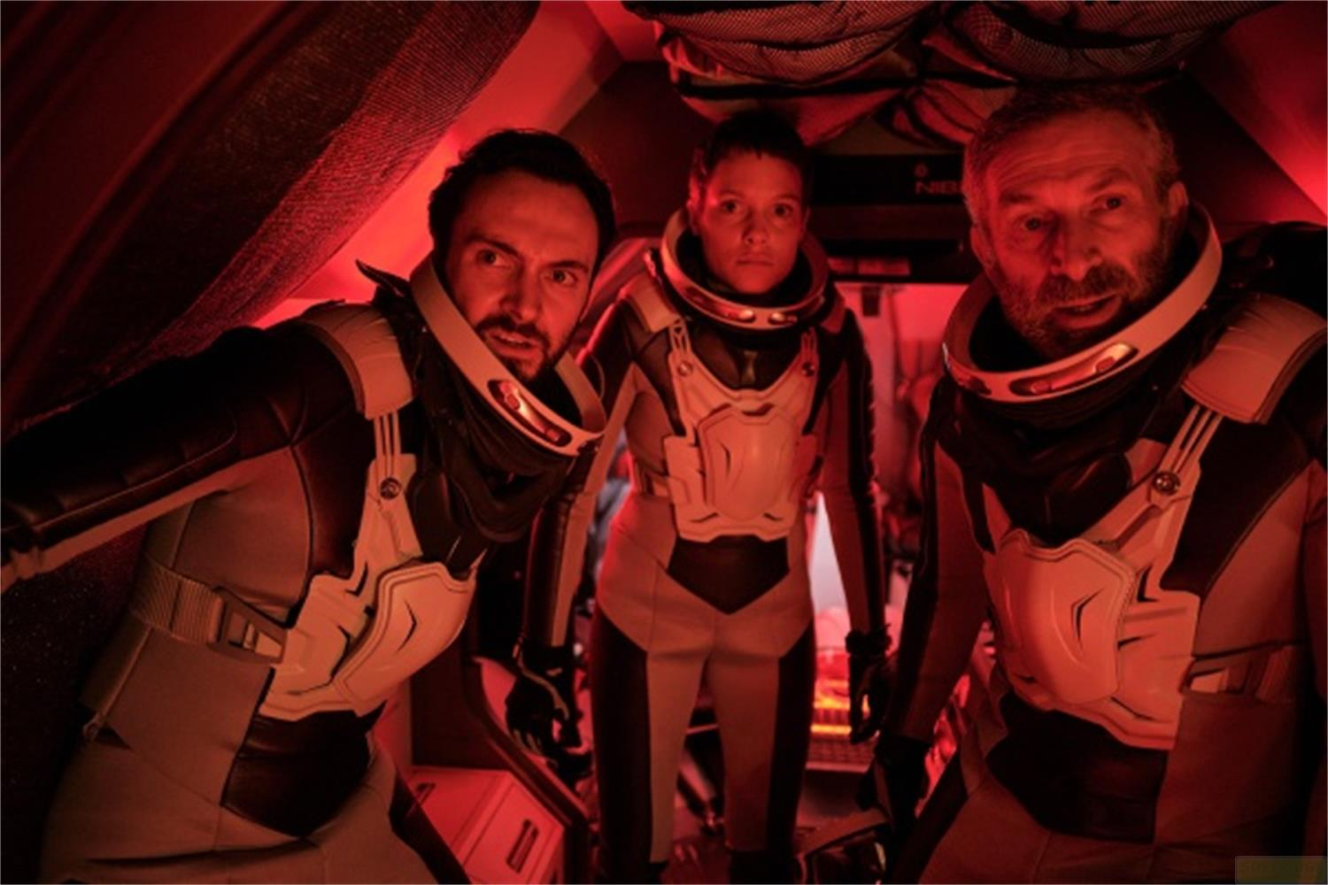 Tre astronauti in tute bianche e grigio scuro sono nella luce rossa, guardando avanti.
