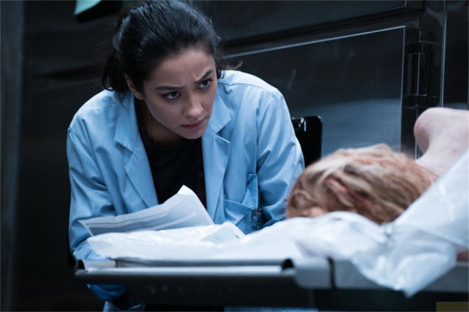 Una donna con camice azzurro si china su un corpo su un tavolo.