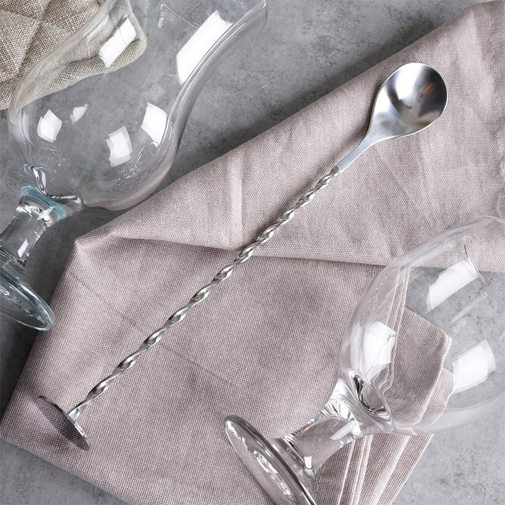 Una cuchara de bar plateada con mango retorcido yace sobre un paño beige junto a dos vasos vacíos.