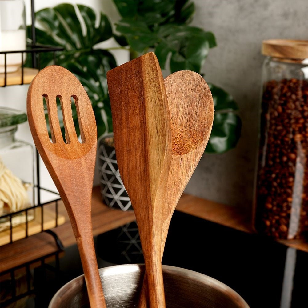 Cucharas y espátula de madera en recipiente metálico, con hojas verdes y tarro al fondo.