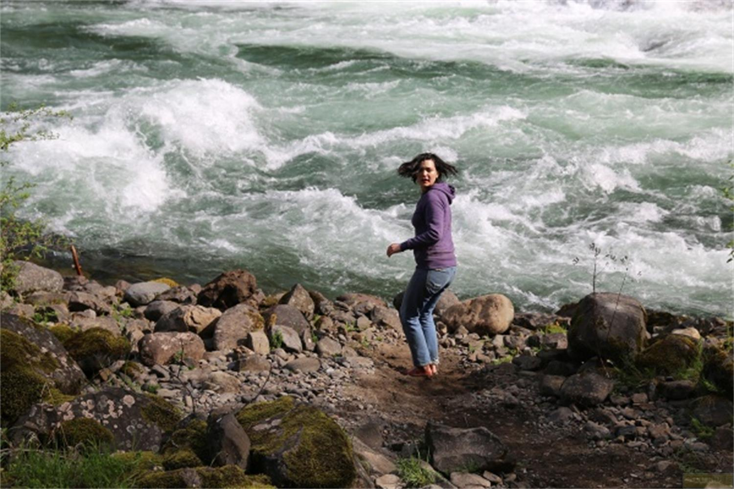 Donna con felpa viola su riva rocciosa vicino a fiume impetuoso.