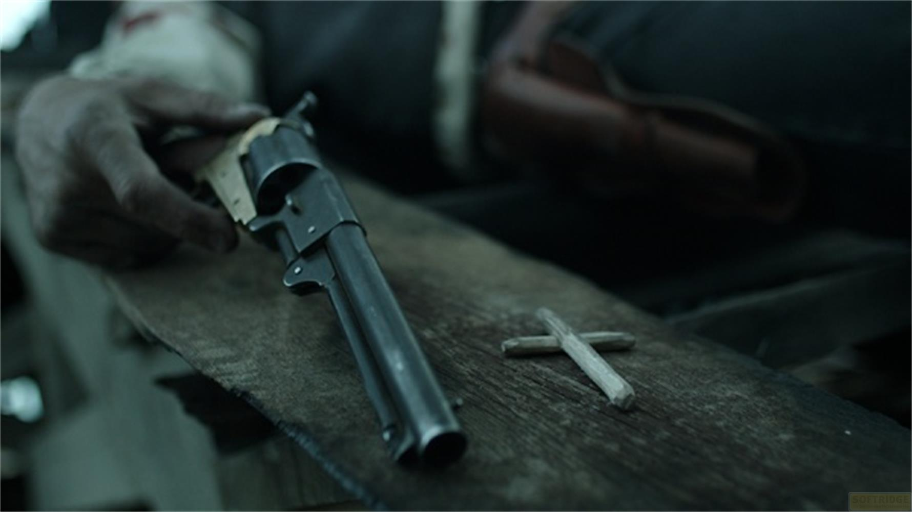 Una mano tiene un revolver su una superficie di legno accanto a una piccola croce di legno.