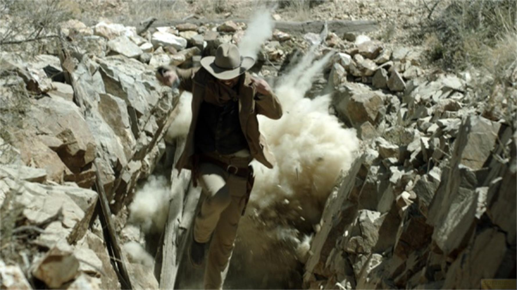Uomo cappello cowboy corre tra rocce, esplosione dietro.