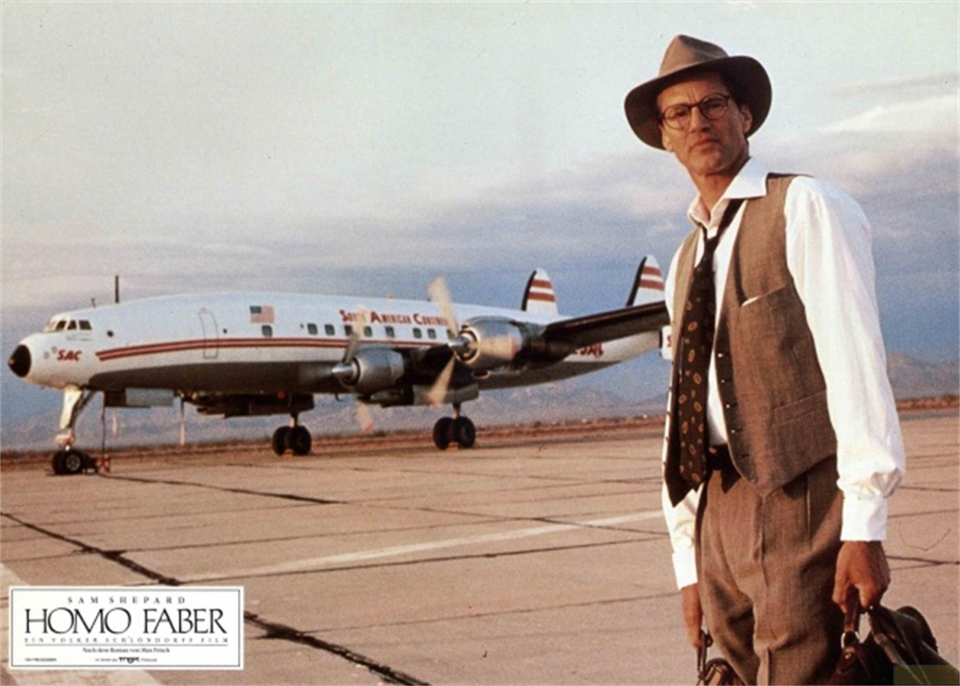 Uomo con cappello e completo in piedi sulla pista accanto a un aereo.