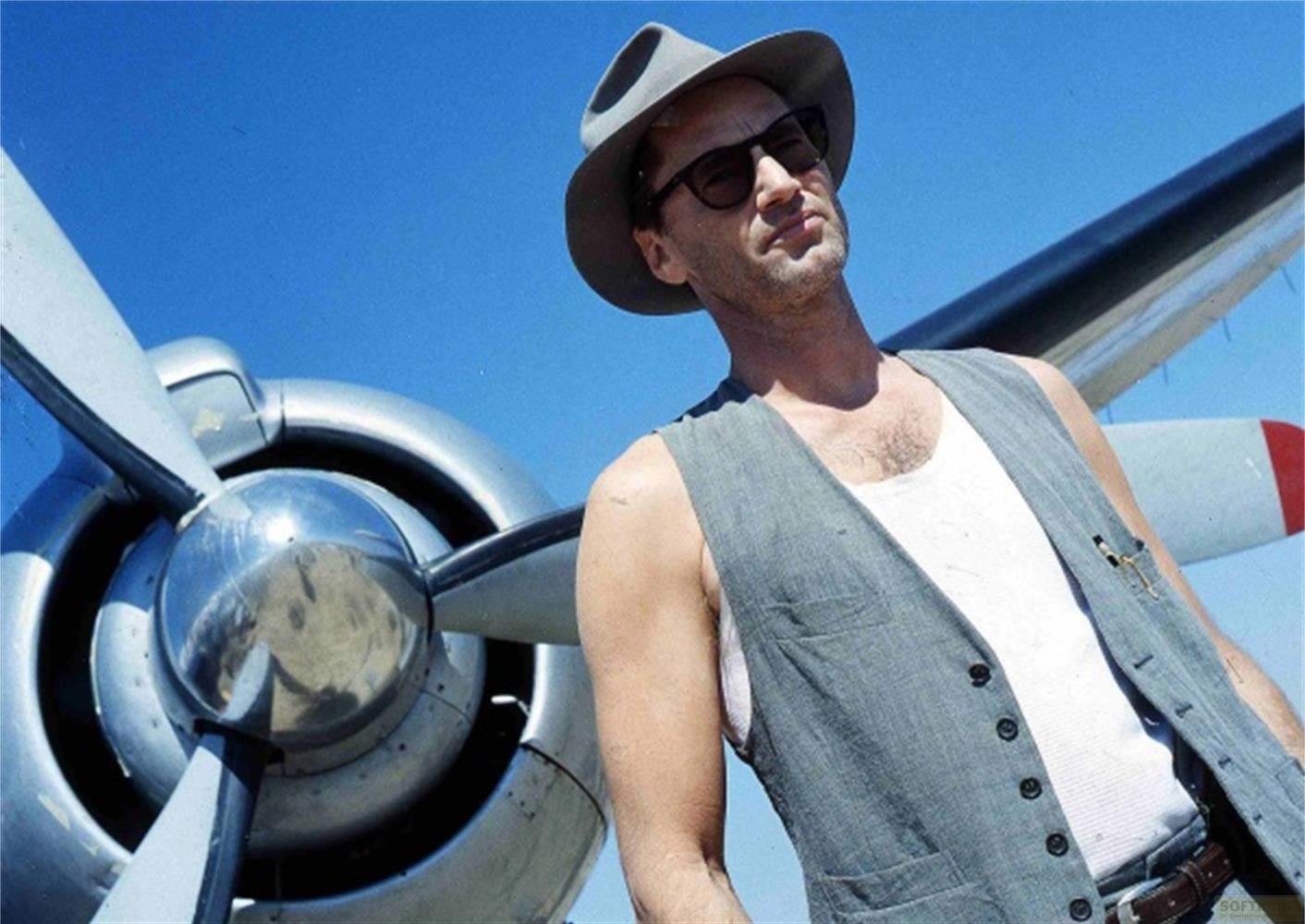 Uomo con cappello e gilet davanti all'elica di un aereo sotto il cielo.
