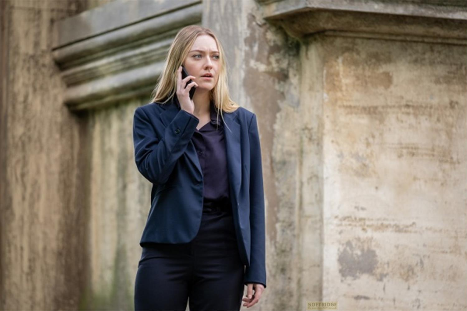Una donna in tailleur blu parla al cellulare all'aperto, contro un muro di pietra.