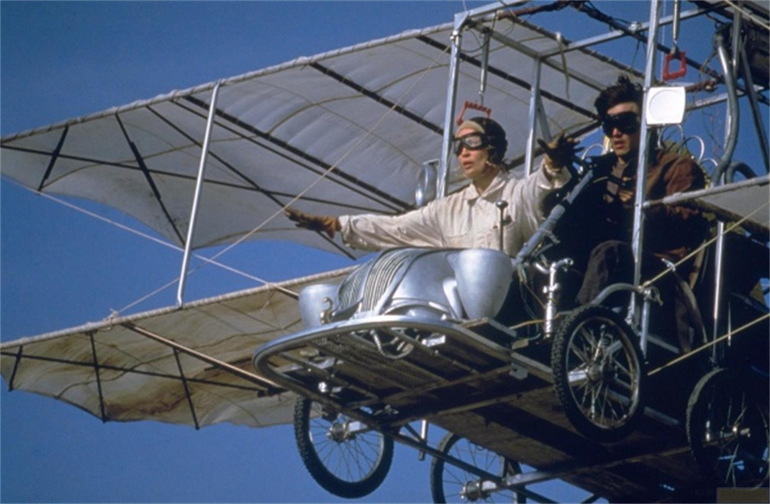 Due piloti in un biplano con frontale d'auto, volano nel cielo azzurro.
