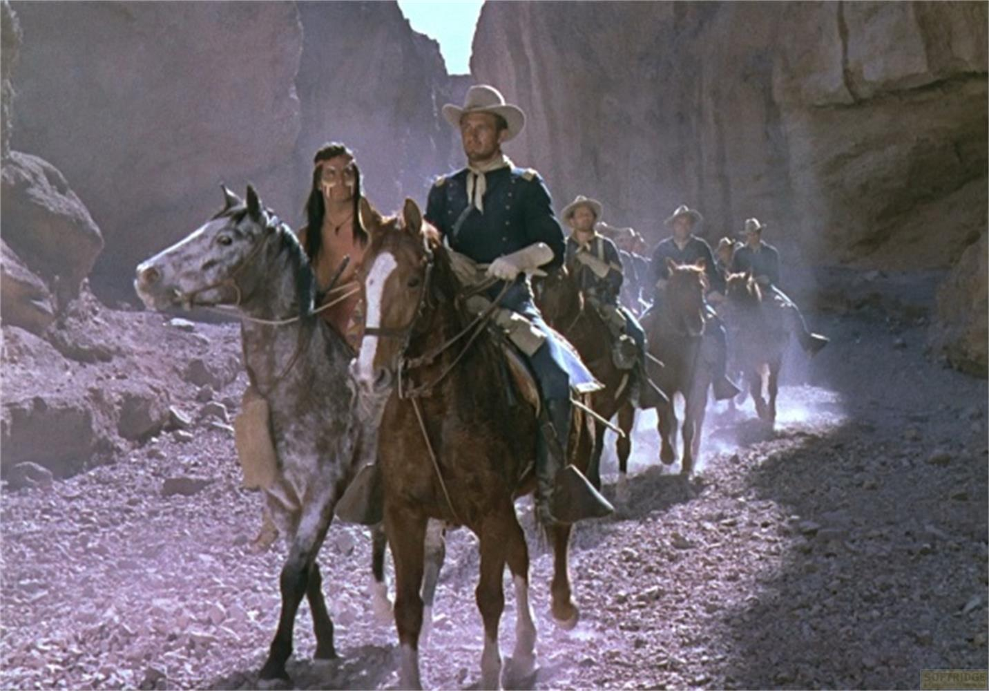 Soldati e un nativo americano cavalcano in un canyon. L'uomo davanti indossa un cappello da cowboy e un'uniforme blu.