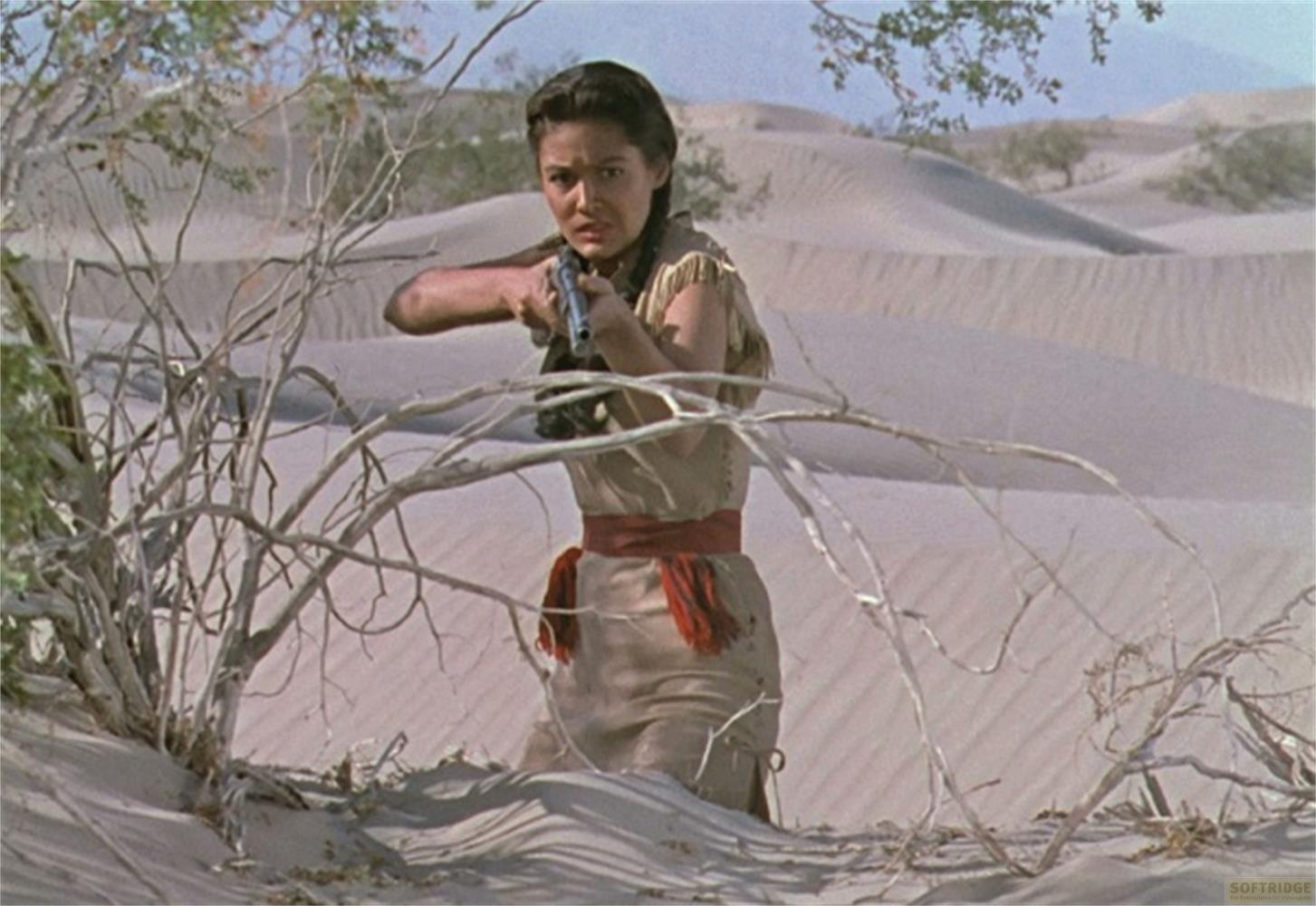 Una donna in abiti desertici mira un fucile tra i rami.