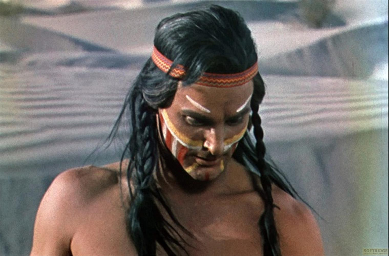 Uomo con pittura di guerra e trecce indossa una fascia su sfondo sabbioso.