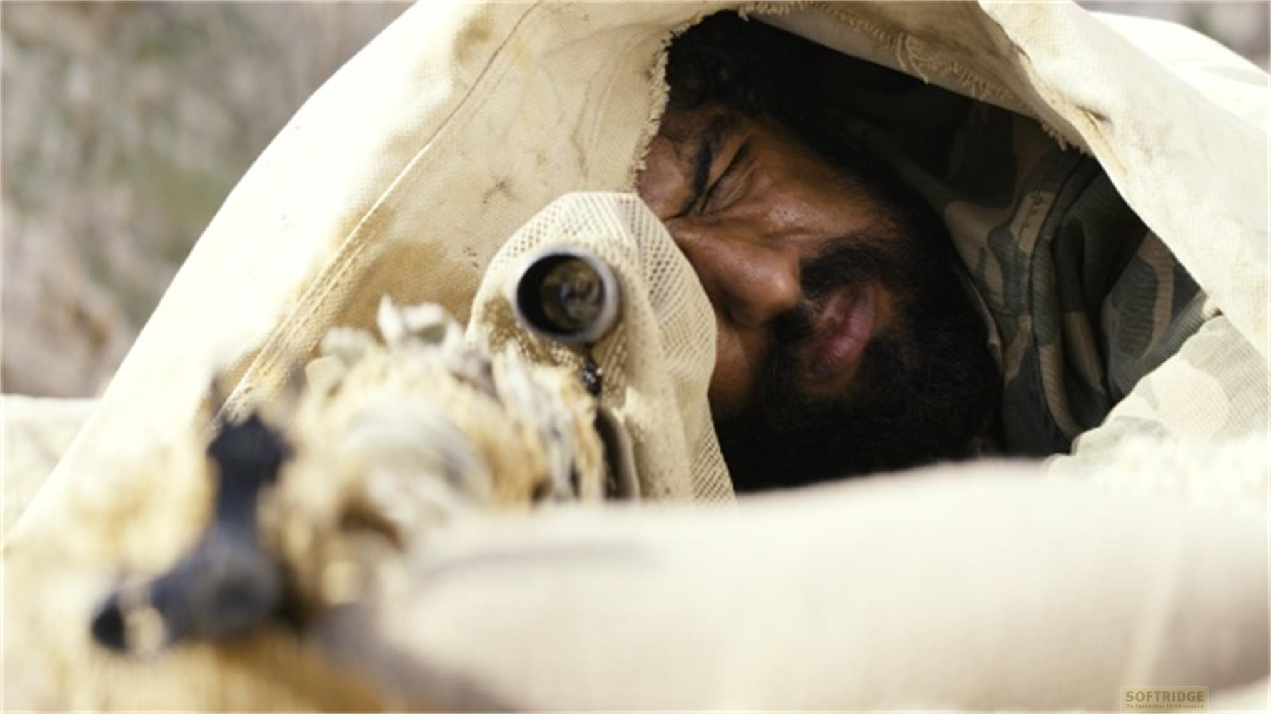 Un uomo con barba e abiti mimetici giace sotto una coperta, guardando attraverso un fucile.