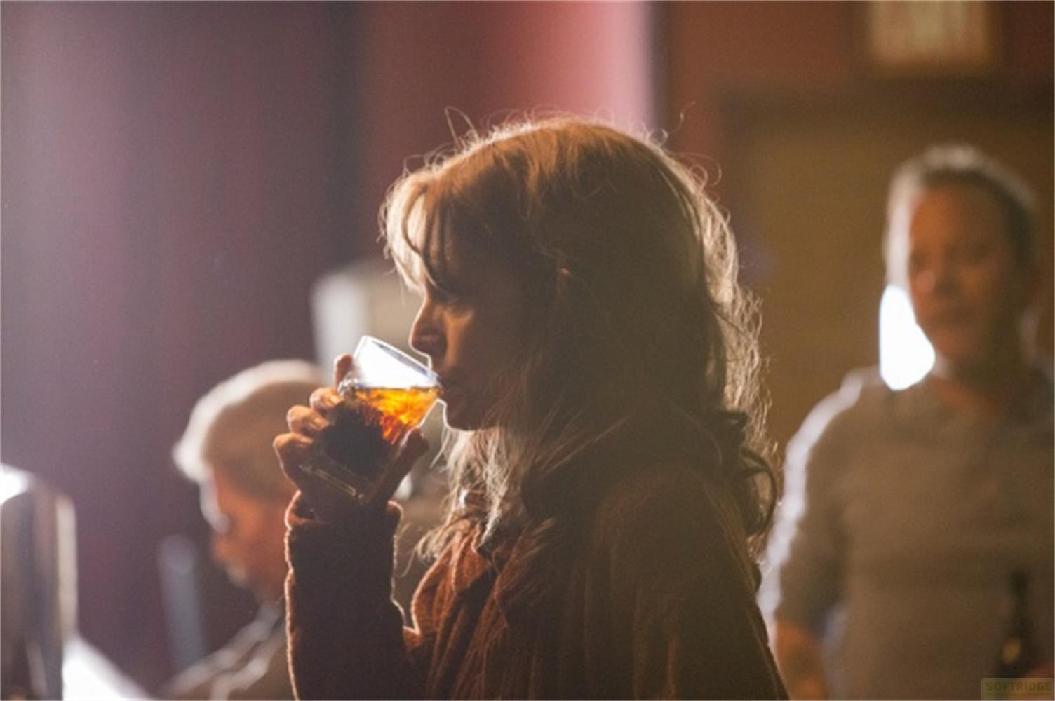 Una donna beve da un bicchiere di liquido scuro. Un'altra persona è sfocata sullo sfondo.