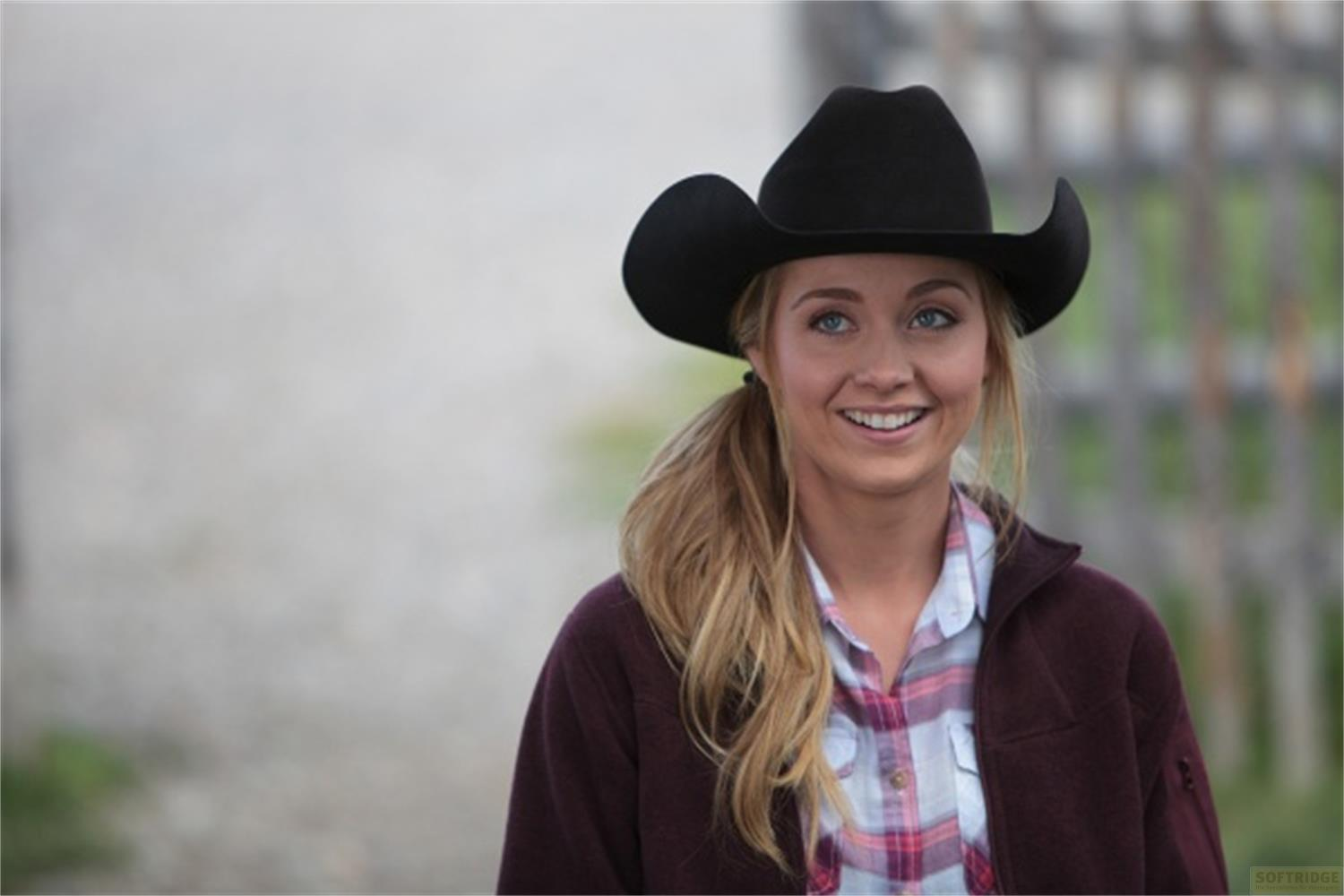 Una giovane donna dai capelli biondi indossa un cappello da cowboy nero, camicia a quadri e giacca bordeaux.
