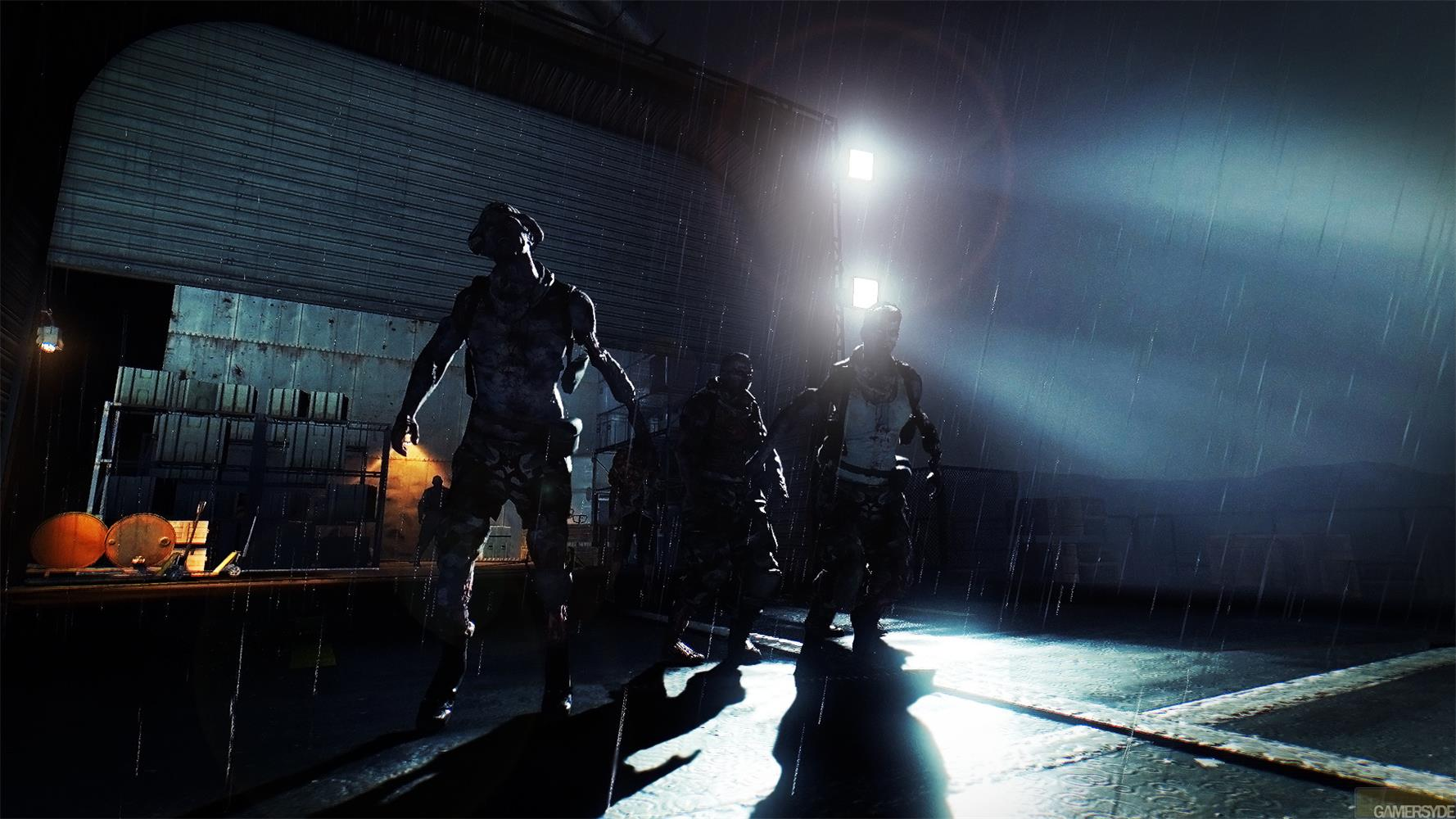 Des zombies sous la pluie devant un hangar, éclairés par des projecteurs.