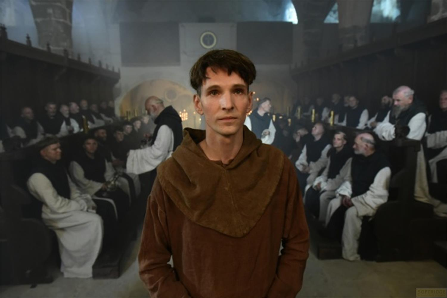 Un giovane con veste da monaco marrone davanti a un gruppo di monaci in un monastero.