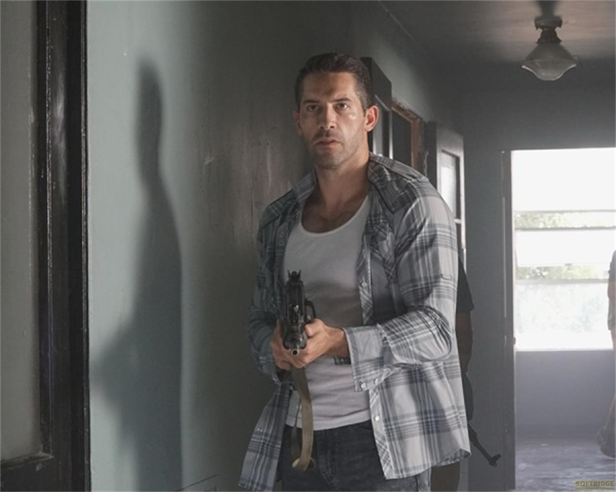 Un uomo con un fucile si trova in un corridoio buio, indossa una camicia a quadri.