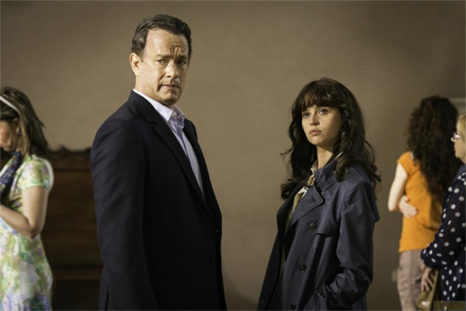 Tom Hanks en costume sombre regarde à gauche, une femme en manteau bleu à ses côtés.