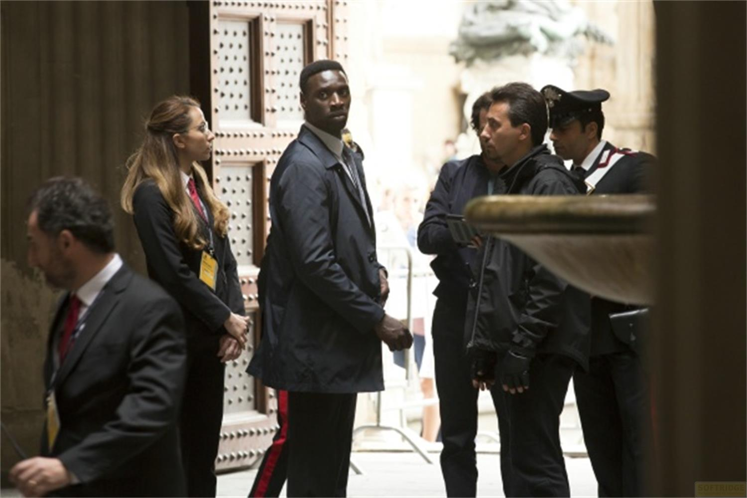 Plusieurs personnes en tenue sombre se tiennent devant une porte. Un homme en uniforme est visible.
