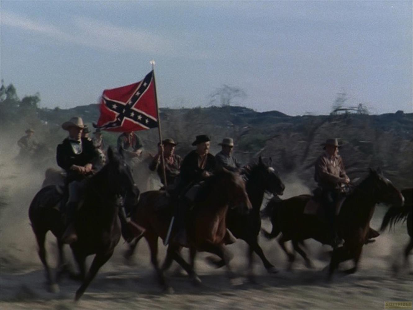 Des cavaliers avec un drapeau confédéré galopent dans un terrain poussiéreux.
