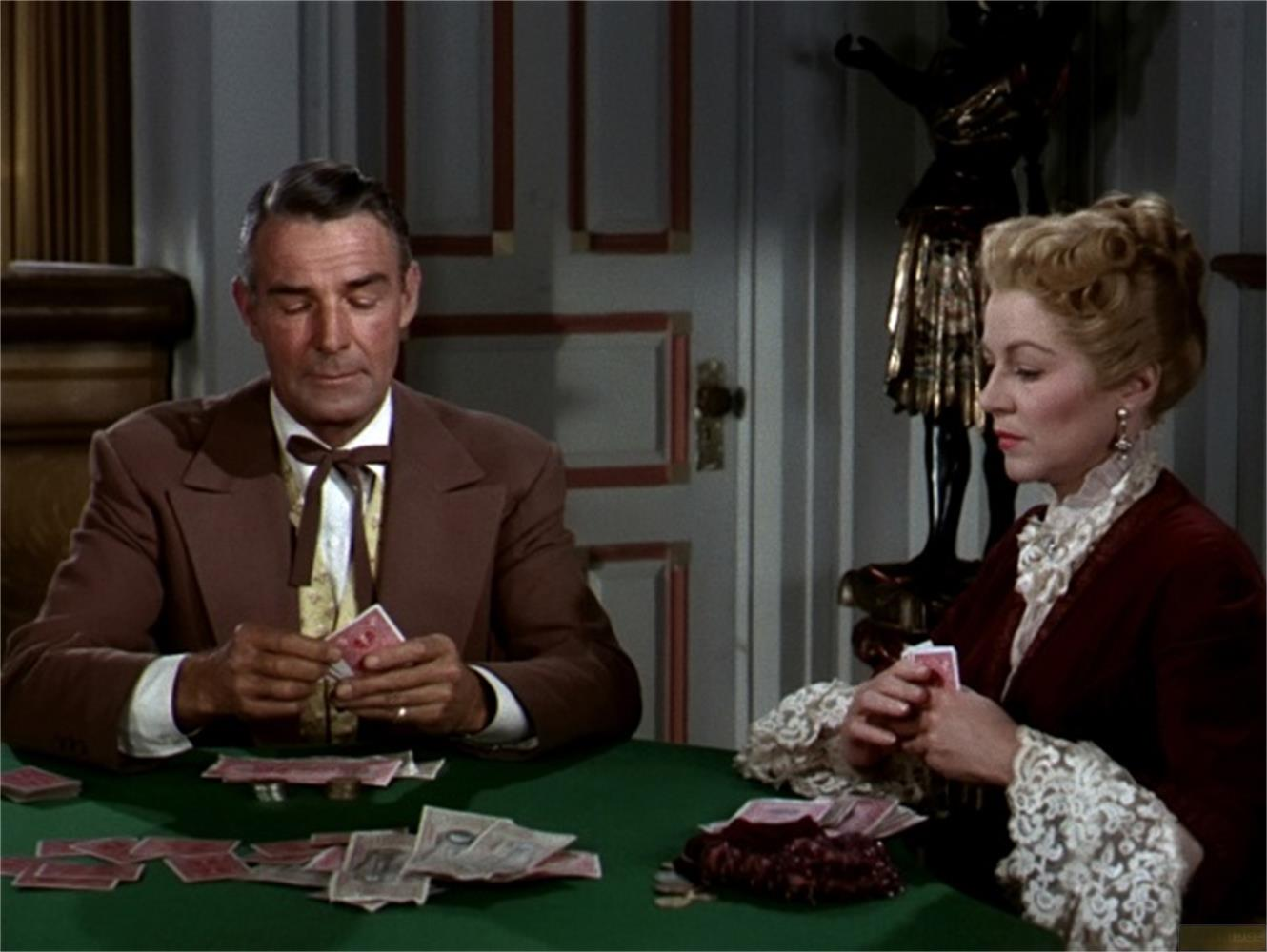 Un homme et une femme assis à une table jouent aux cartes. L'homme porte un costume marron et la femme une robe rouge.