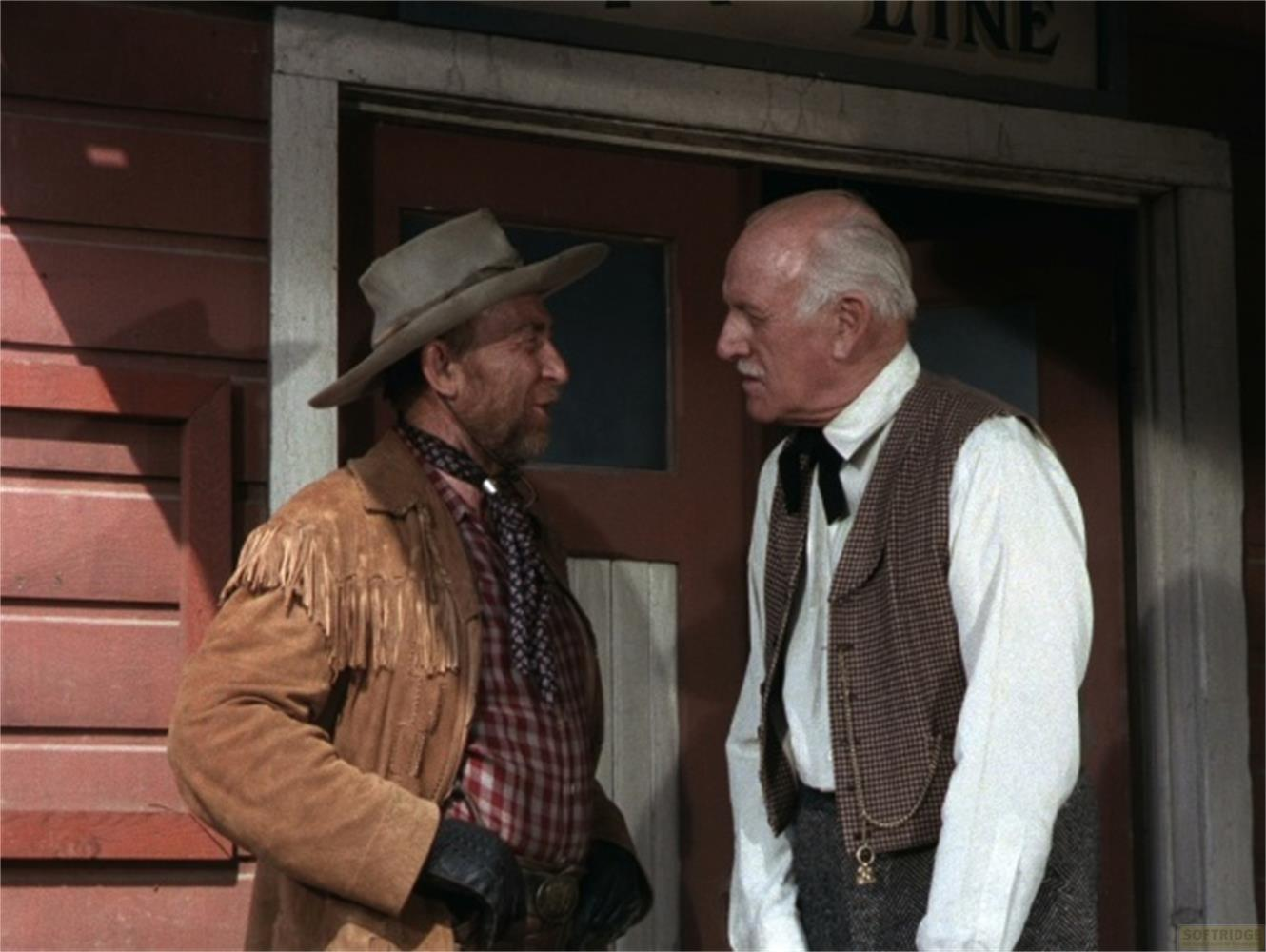 Deux hommes devant un bâtiment en bois. L'un porte un chapeau et une veste à franges.