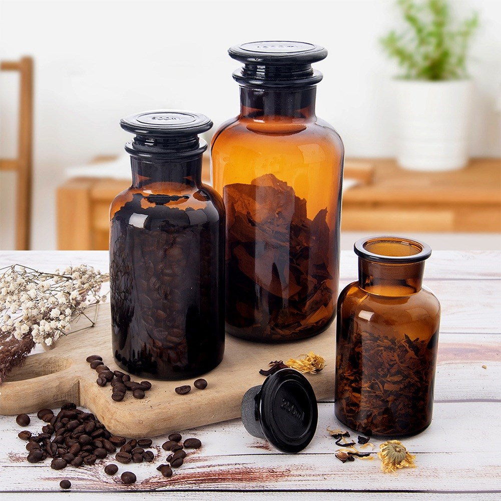 Tres frascos de vidrio ámbar con tapas negras, llenos de granos de café y hierbas.