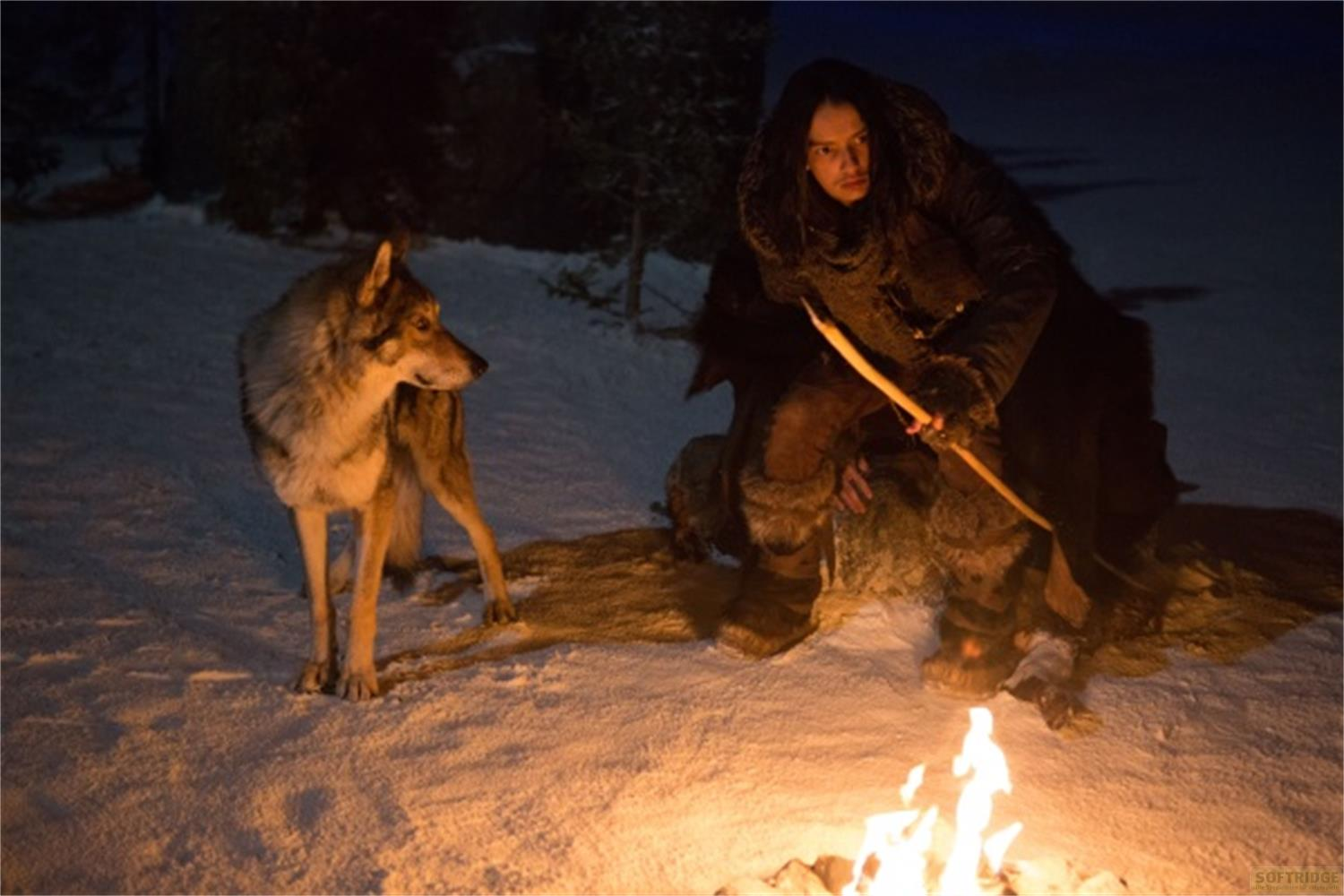 Un uomo in abiti invernali e un lupo siedono vicino a un falò nella neve.