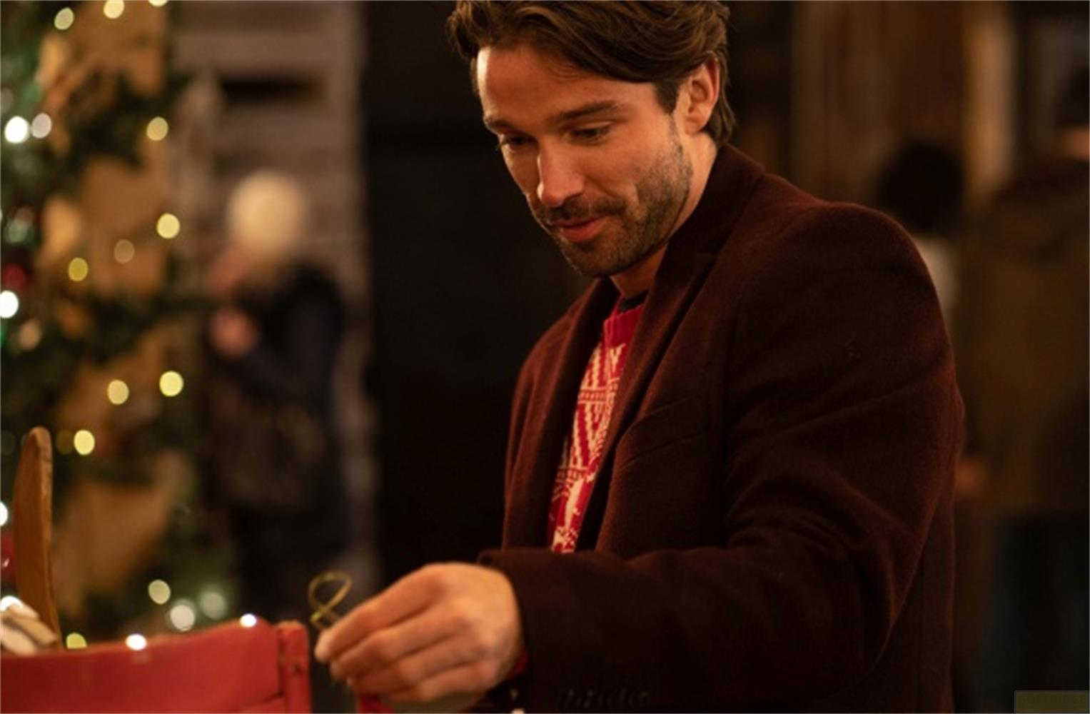 Uomo con maglione natalizio e cappotto marrone crea qualcosa vicino a un albero di Natale.