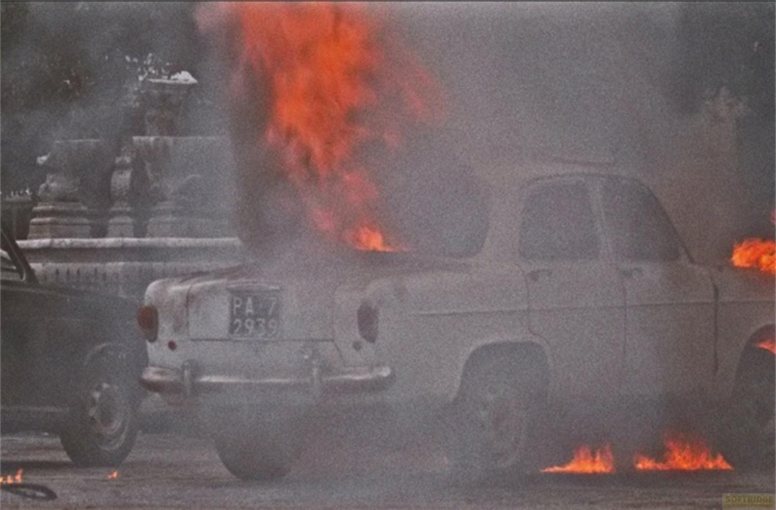 Un'auto d'epoca bianca è in fiamme in una strada, con fumo che sale.
