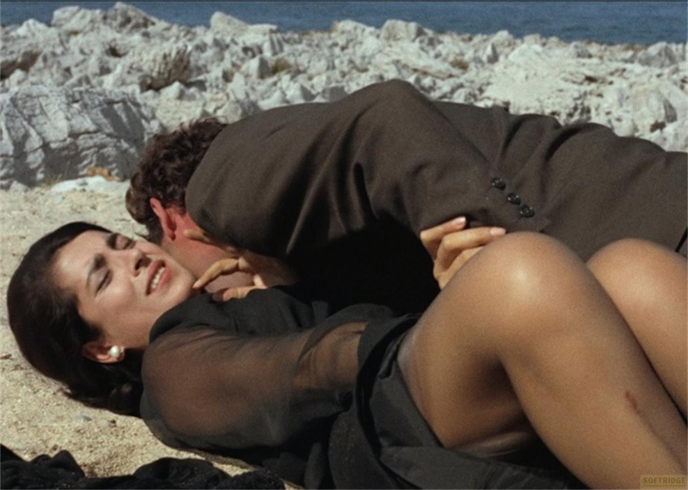 Un uomo in abito marrone bacia una donna sdraiata su una spiaggia sabbiosa.
