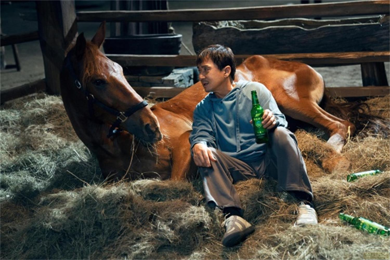 Un uomo siede nel fieno accanto a un cavallo marrone, tenendo una bottiglia verde.