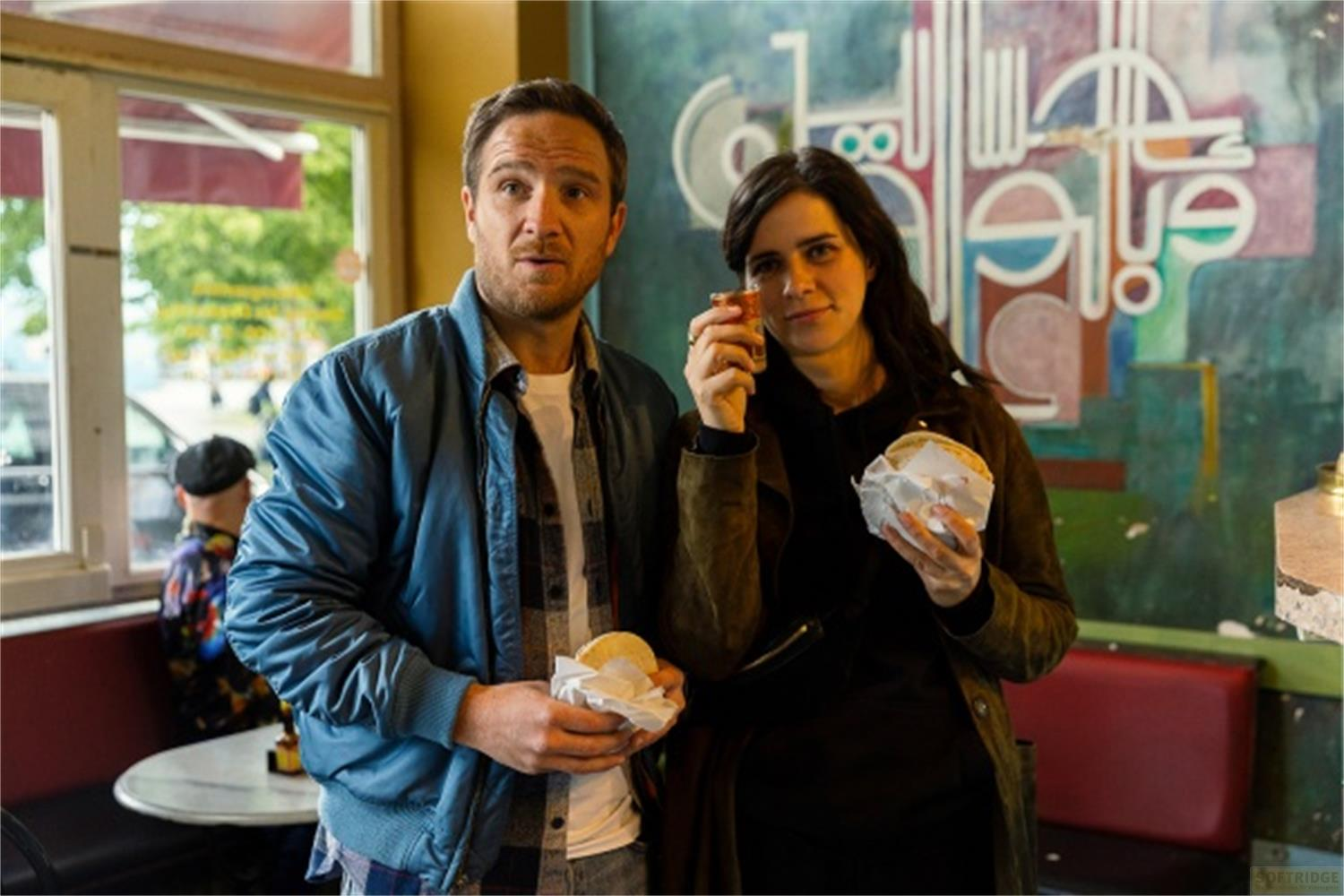 Un uomo e una donna tengono degli snack. L'uomo indossa una giacca blu. Lo sfondo presenta un murale.