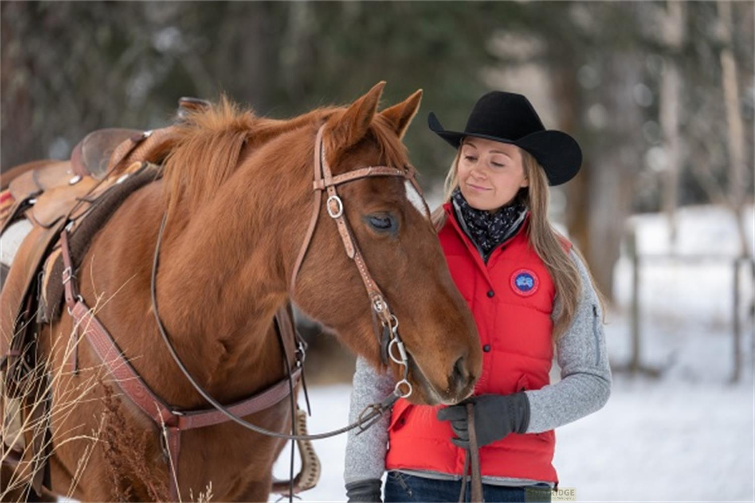Una donna con gilet rosso e cappello nero accarezza un cavallo marrone nella neve.