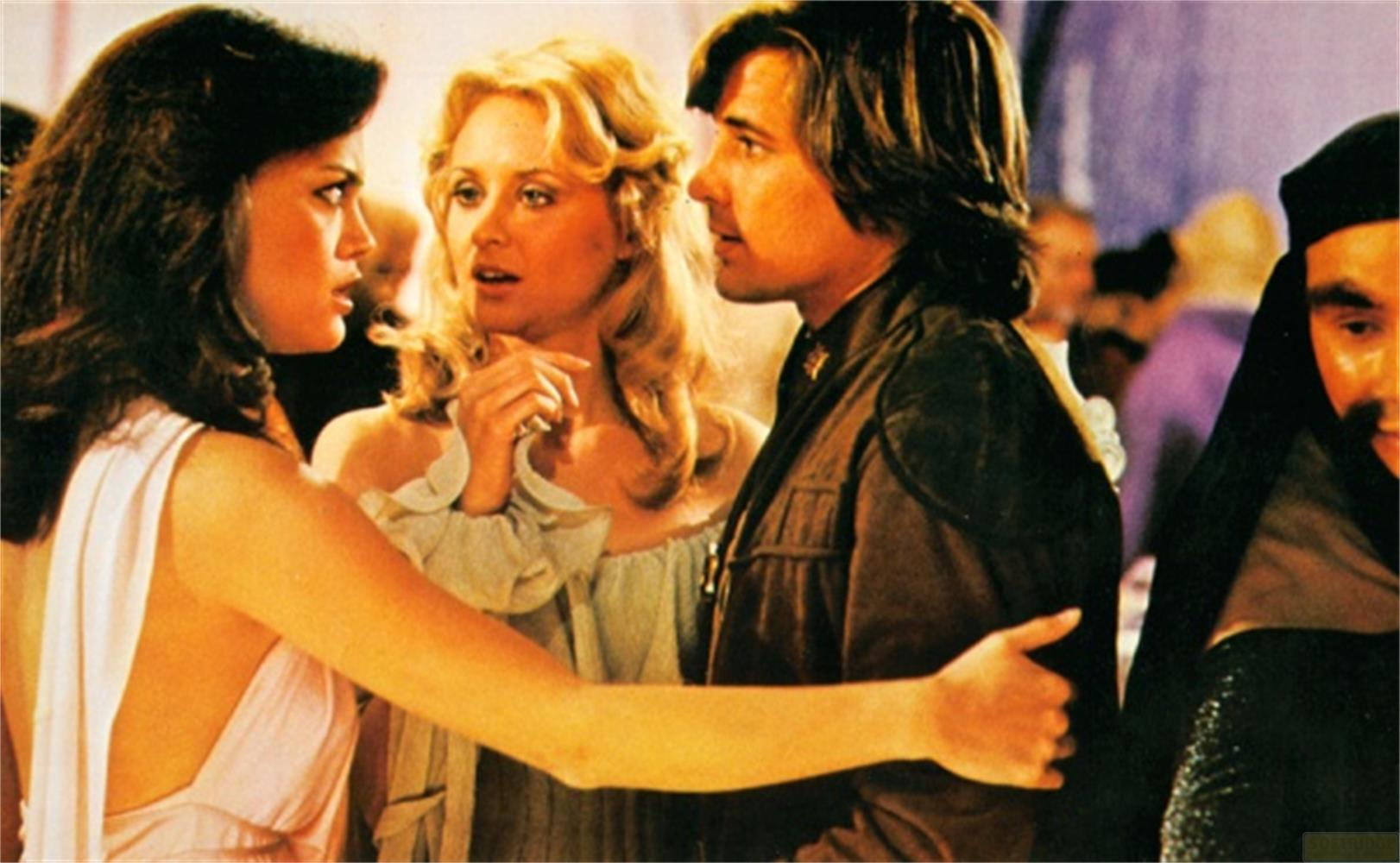 Tre persone in primo piano: donna in rosa, donna in azzurro, uomo in giacca marrone.