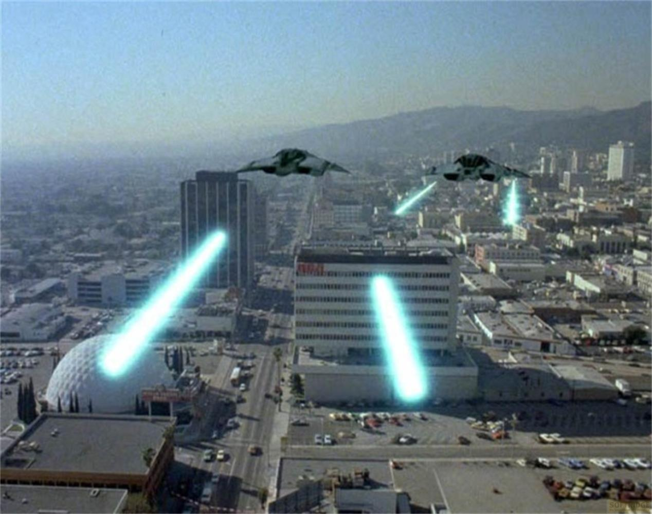 Navi aliene sparano laser blu sullo skyline di una città con edifici e montagne.