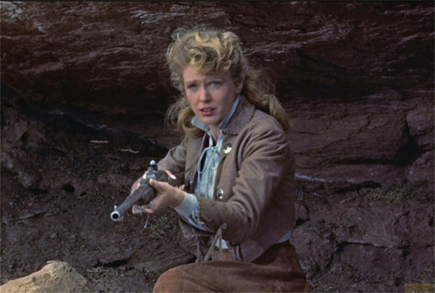 Una donna bionda tiene una pistola in un ambiente roccioso.
