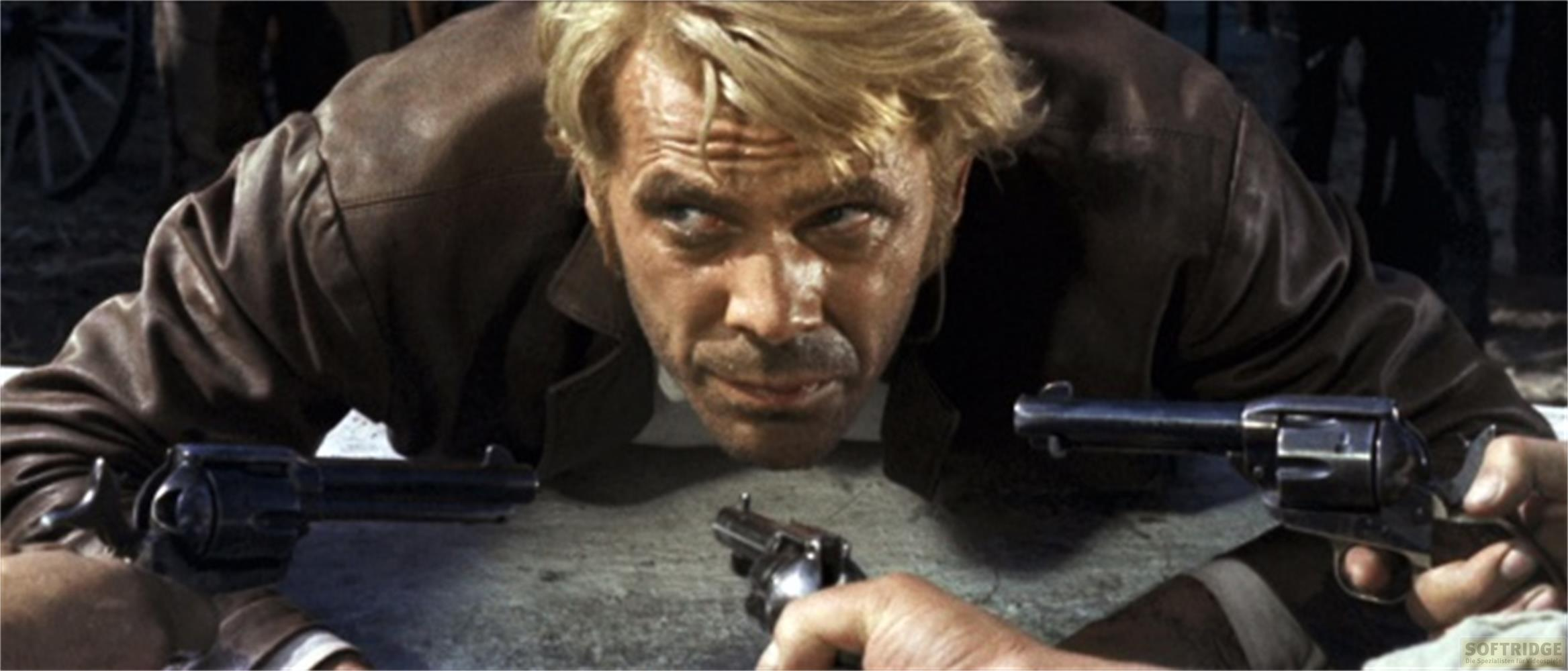 Un uomo biondo con una giacca marrone si china con tre revolver, uno in ogni mano e uno puntato su di lui.