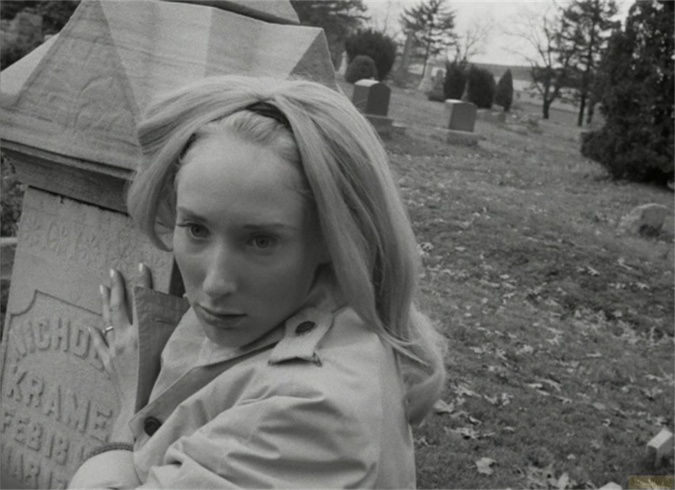 Una donna bionda si appoggia a una lapide in un cimitero.