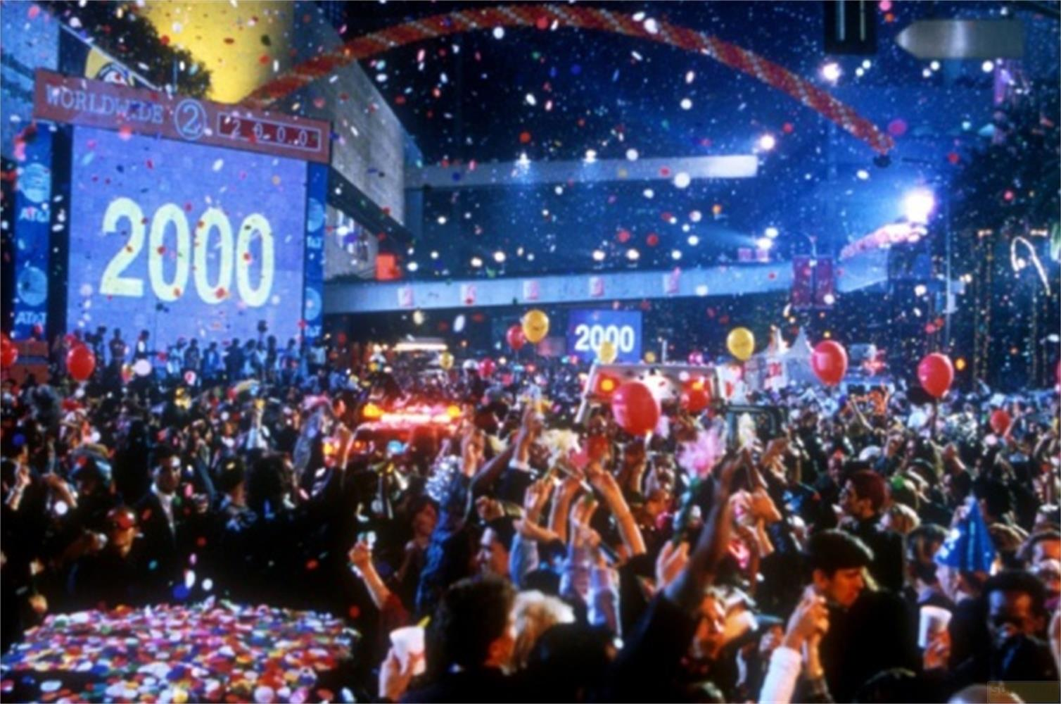 Folla festeggia Capodanno con coriandoli e palloncini davanti a un grande schermo con scritto '2000'.