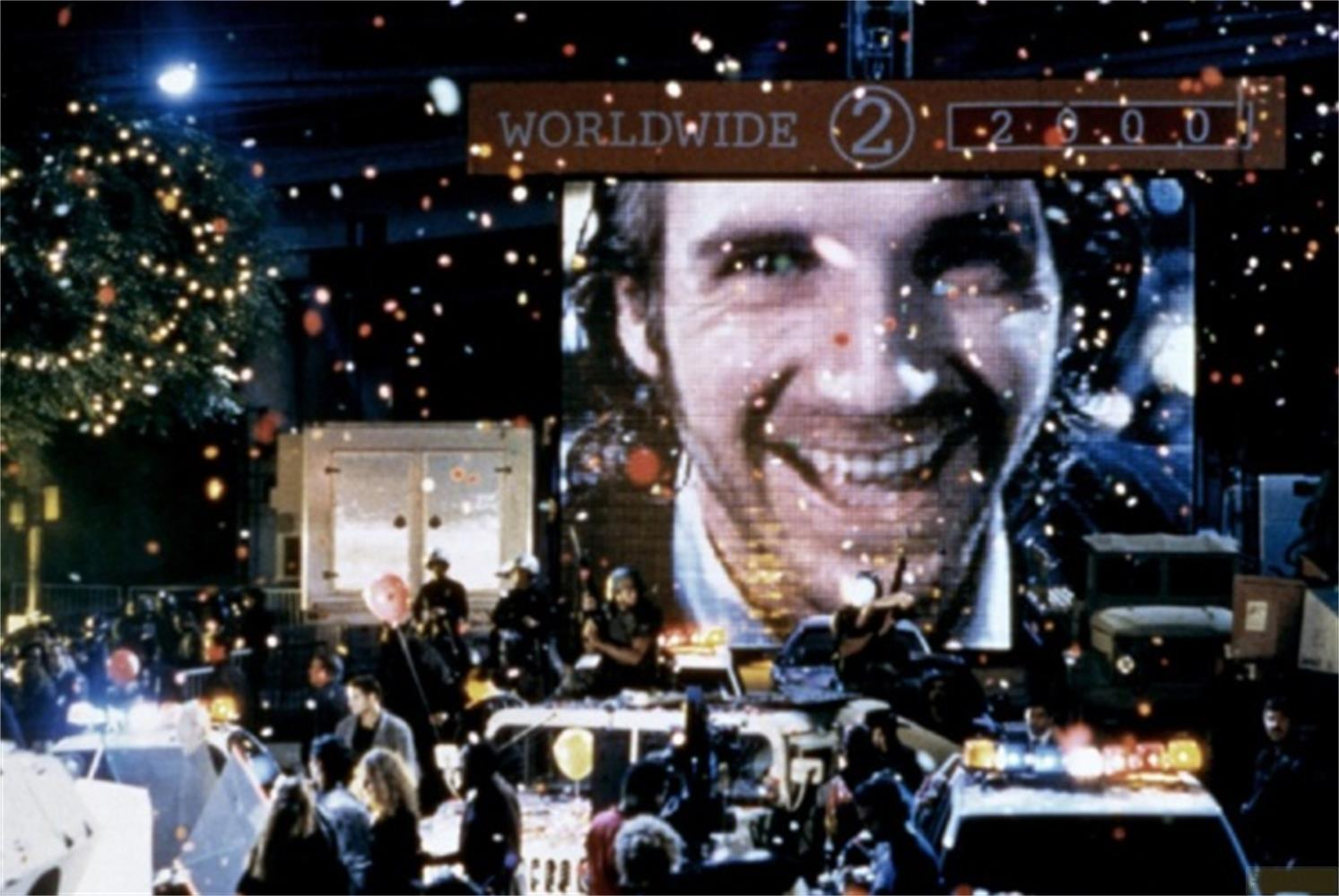 Grande schermo mostra un volto sorridente. Sotto, folla, auto, coriandoli.