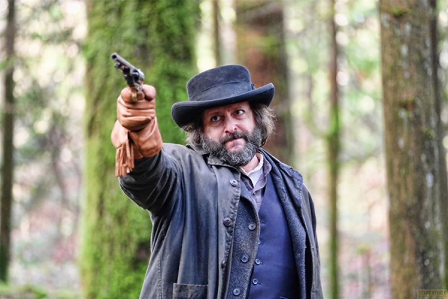 Un uomo con cappello e guanti alza una pistola in un bosco.
