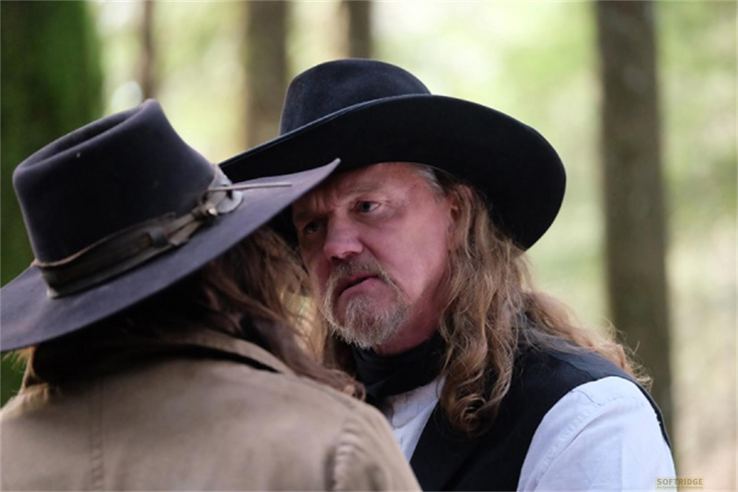 Due uomini con cappelli da cowboy in una foresta. Uno guarda di lato.