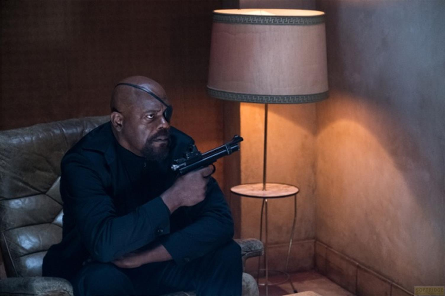 Ein Mann mit Augenklappe sitzt mit Waffe in einem Sessel.
