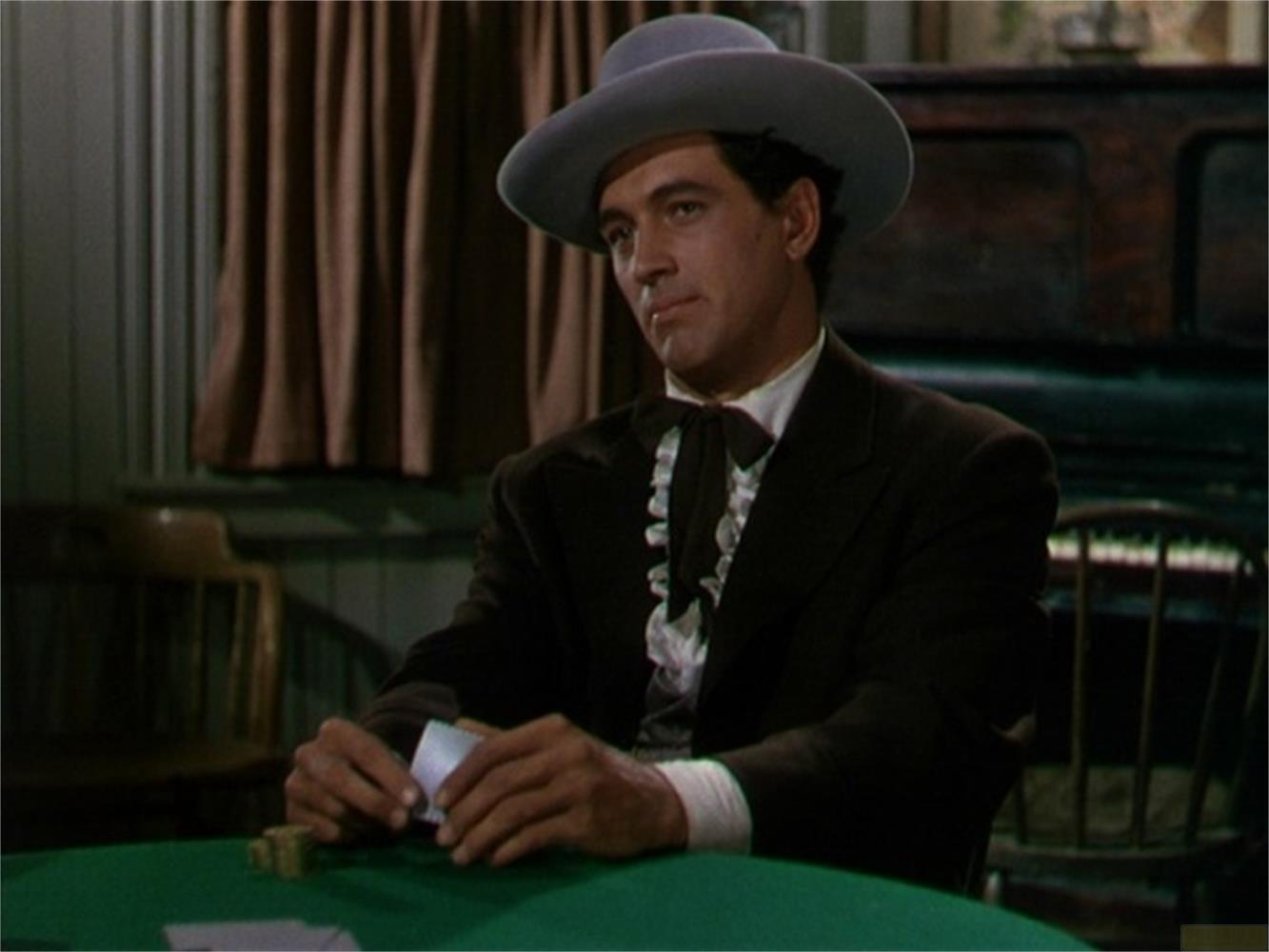 Uomo con cappello gioca a carte a tavolo verde, con sedie di legno sullo sfondo.