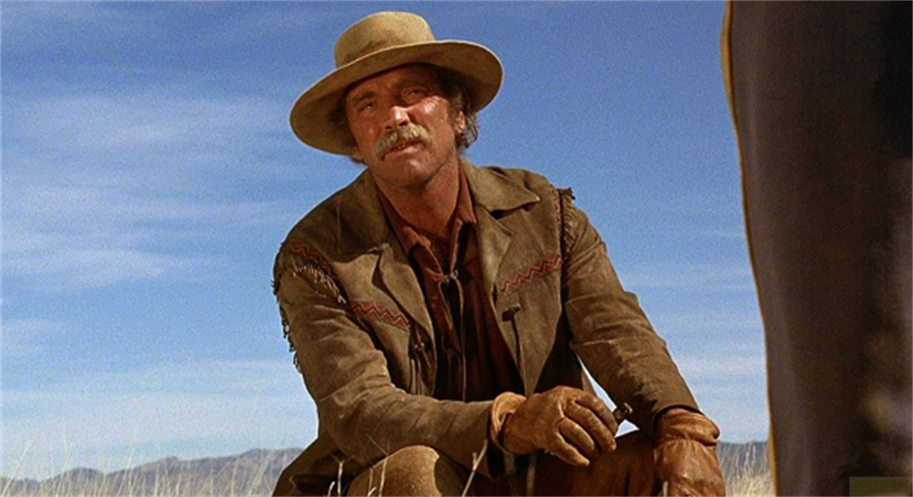 Un uomo con i baffi, cappello e giacca di camoscio si inginocchia all'aperto.