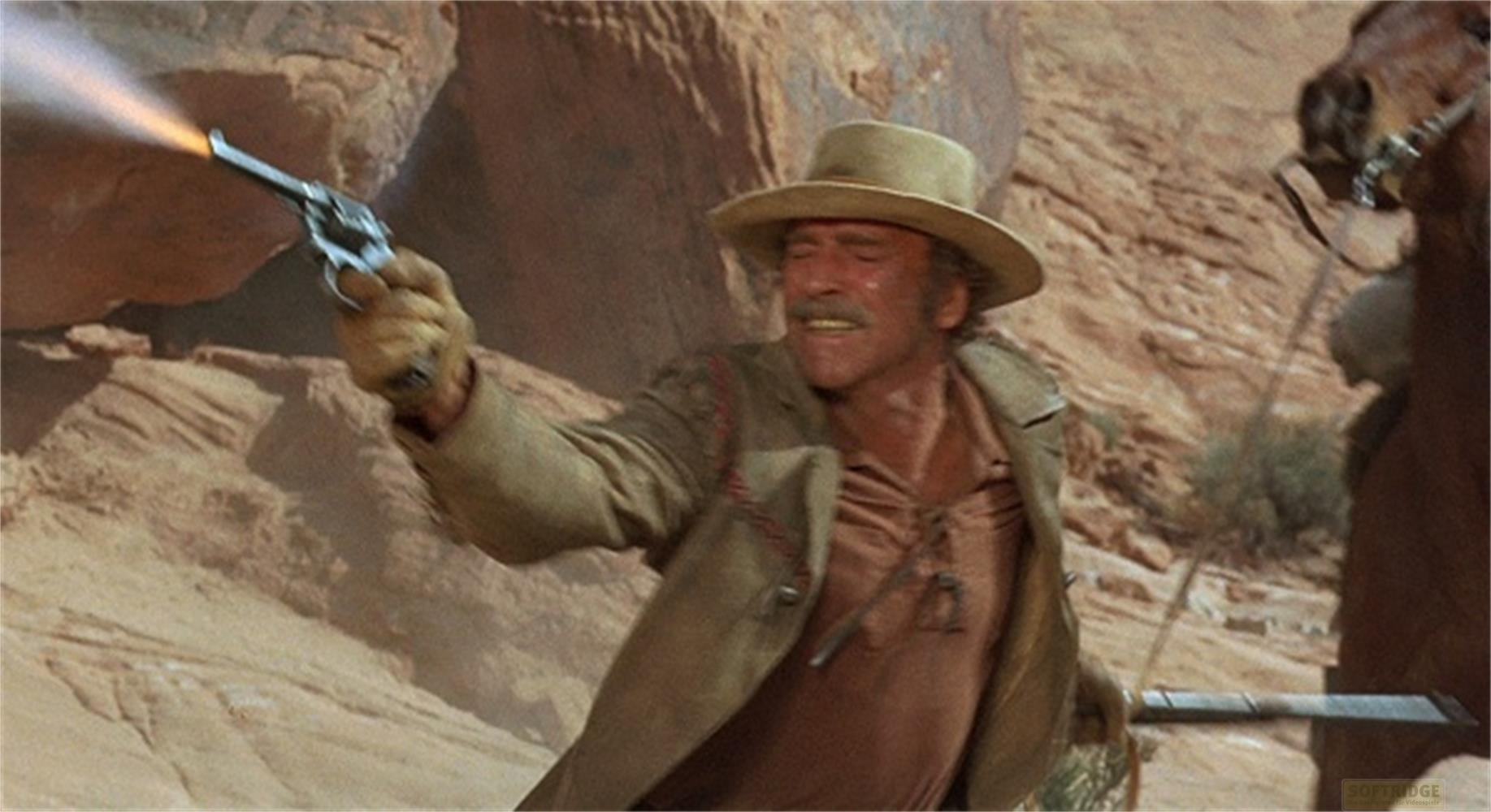 Uomo con cappello western spara pistola, fumo, sfondo roccioso.