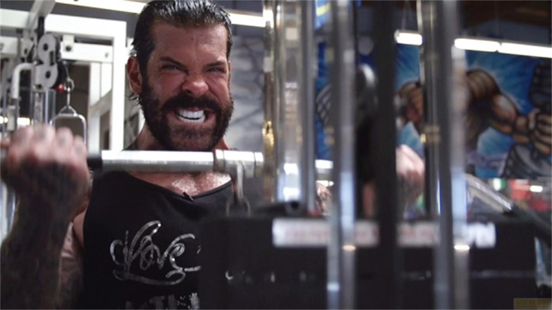 Un uomo con la barba solleva un bilanciere in palestra, il viso contratto.