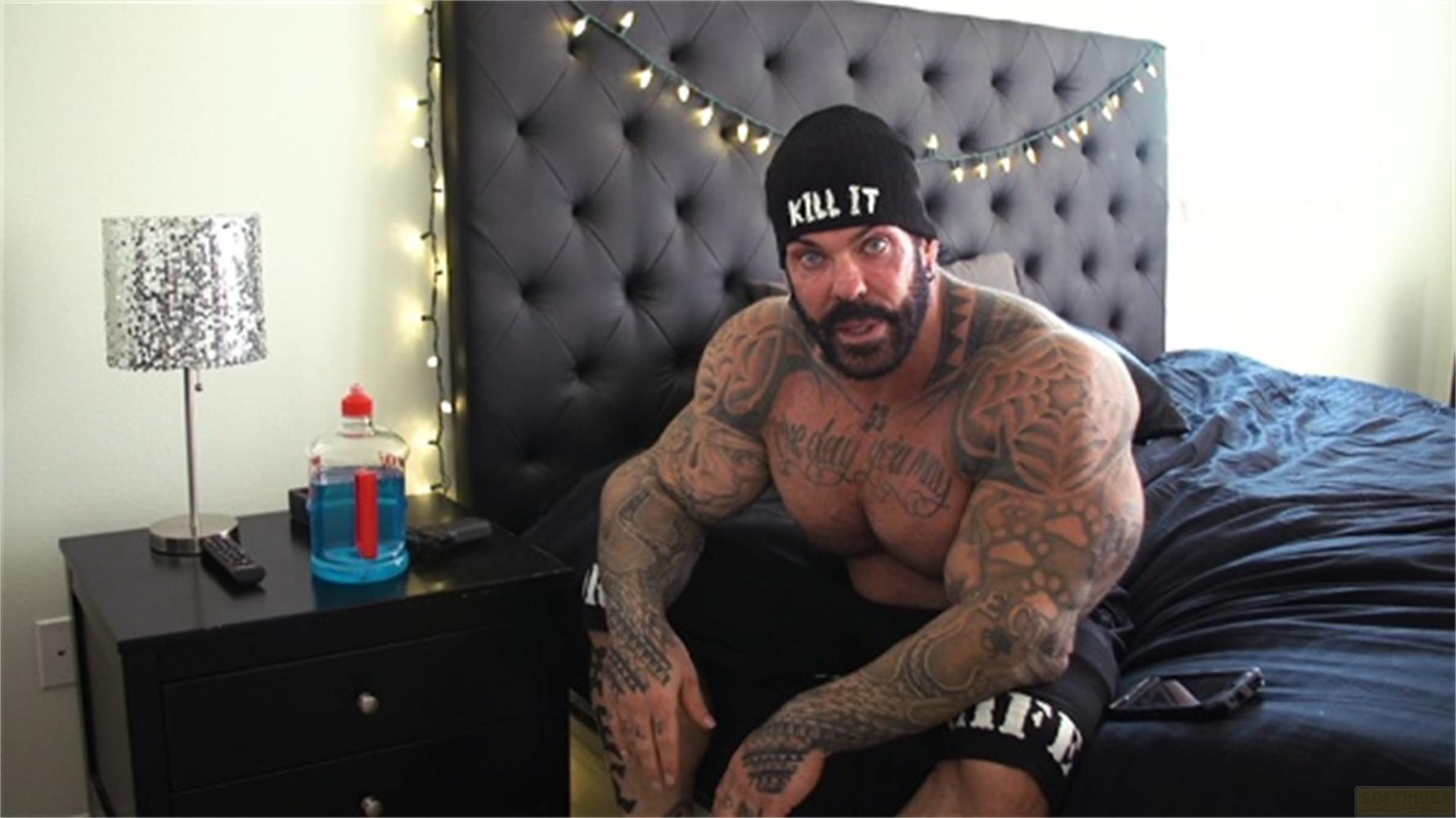 Un uomo muscoloso con tatuaggi è seduto su un letto, indossa un berretto nero.