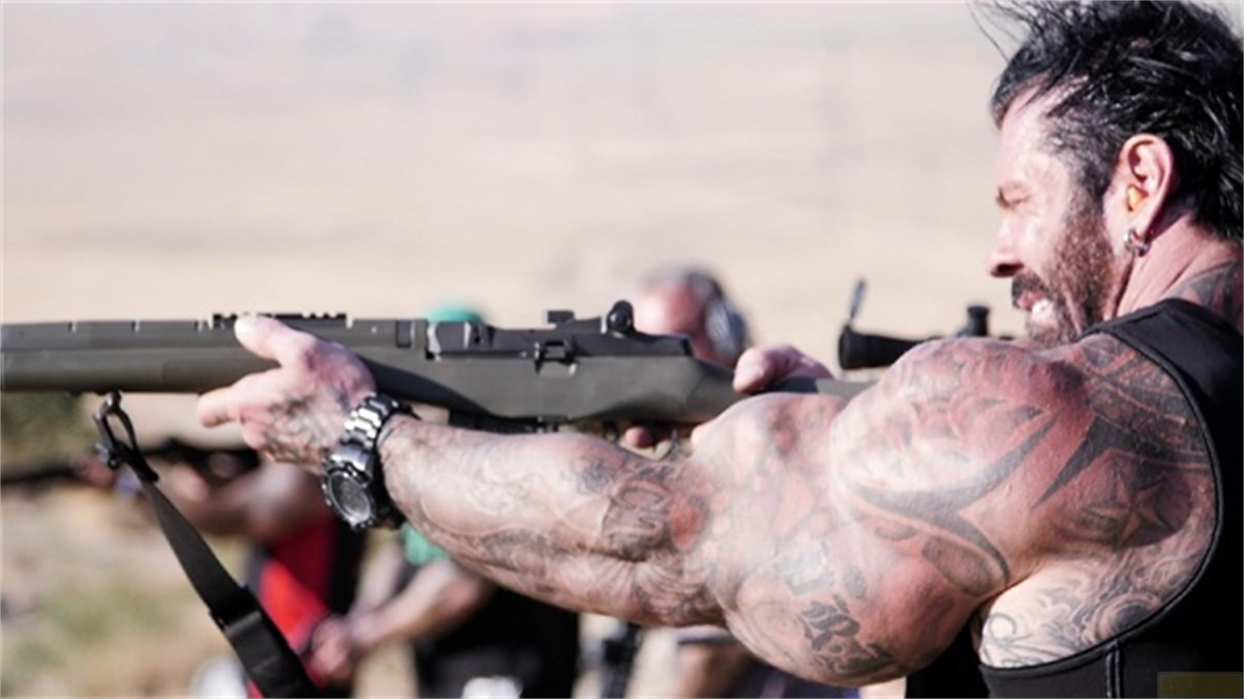 Un uomo tatuato punta un fucile all'aperto in un paesaggio desertico.